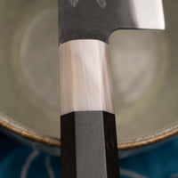 Yoshida Gyuto SUJ-2 Kuro-uchi 210 mm je ručno izrađen kuhinjski nož srednje veličine od izdržljivog SUJ-2 čelika s rustikalnim kuro-uchi završetkom, idealan za razne zadatke i s jedinstvenom gravurom koi šarana.