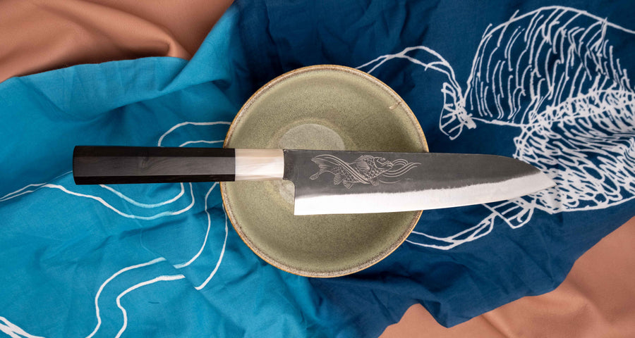 Yoshida Gyuto SUJ-2 Kuro-uchi 210 mm je ručno izrađen kuhinjski nož srednje veličine od izdržljivog SUJ-2 čelika s rustikalnim kuro-uchi završetkom, idealan za razne zadatke i s jedinstvenom gravurom koi šarana.