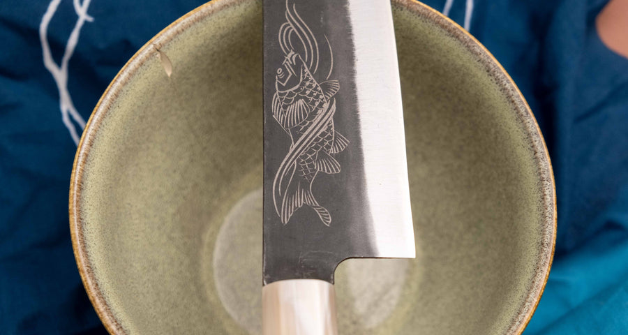 Yoshida Gyuto SUJ-2 Kuro-uchi 210 mm je ručno izrađen kuhinjski nož srednje veličine od izdržljivog SUJ-2 čelika s rustikalnim kuro-uchi završetkom, idealan za razne zadatke i s jedinstvenom gravurom koi šarana.