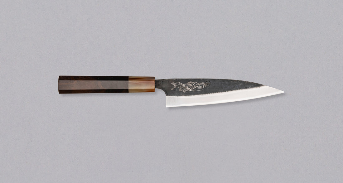 Yoshida Petty SUJ-2 Kuro-uchi 145 mm je ručno izrađen kuhinjski nož srednje veličine od izdržljivog SUJ-2 čelika s kuro-uchi završetkom, koji osigurava rustikalan izgled i iznimnu postojanost oštrine. Opremljen ručkom od ebanovine za siguran zahvat i jedinstvenom gravurom koi šarana, idealan je za razna rezanja i sjeckanja te savršen kao prvi japanski nož ili kao poklon za ljubitelje japanske kuhinje.